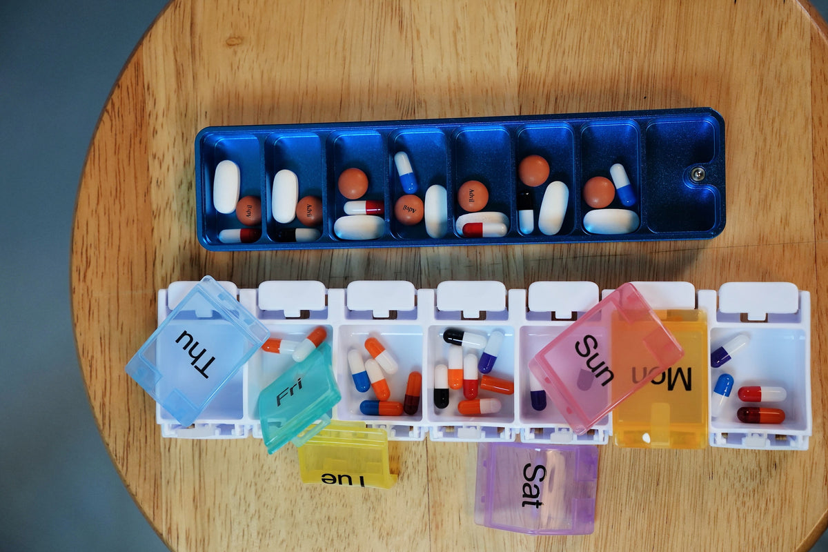 Plastic weekly pill organizer with lids hanging open next to an Ikigai aluminum case