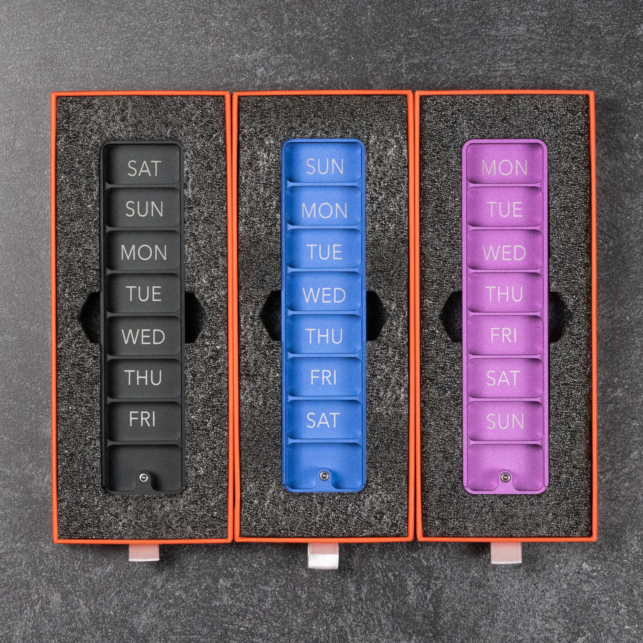 Three weekly pill organizers in black, blue, and purple with labeled compartments for each day.