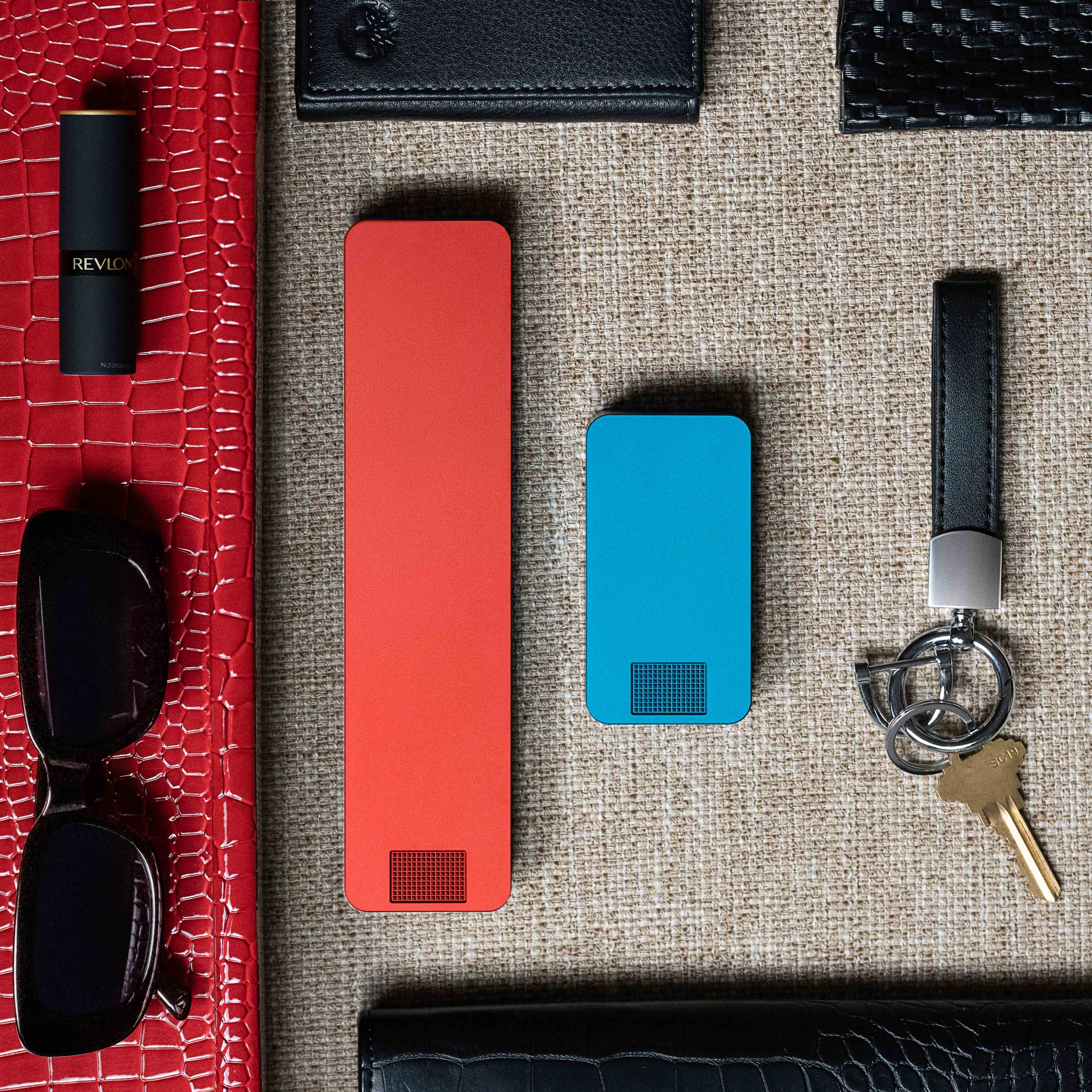 Everyday items including lipstick, keys, two smartphones, sunglasses, wallets, and a cord on an orange and beige background.