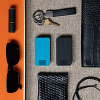 One Bahama Blue and One Matte Black Travel Pill Case laying next to a coin purse, women's sunglasses, lipstick, wallet and keys.